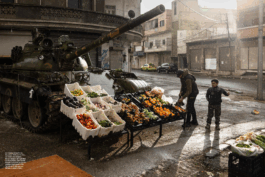 Doppelseite aus den „Fotos für die Pressefreiheit 2025“: Ein syrischer Straßenhändler verkauft frisches Gemüse und Obst auf einem provisorischen Marktstand, der auf einem verlassenen Militärpanzer aufgebaut ist. Ein Junge beobachtet die Szene. Im Hintergrund leerstehende Häuser bei Nieselregen.