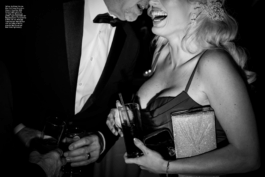 Doppelseite aus den „Fotos für die Pressefreiheit 2025“ mit einem Foto von Mark Peterson: Ein Mann und eine Frau in Abendkleidung lachen ausgelassen auf einer Gala-Veranstaltung. Die Frau trägt ein Cocktailkleid und hält ein Glas in der Hand, der Mann flüstert ihr etwas zu. Die Szene thematisiert die Nähe zwischen Politik und High Society.